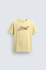 Camiseta de corte relajado color amarillo con efecto lavado. Presenta cuello redondo, mangas cortas con dobladillo y un estampado frontal con texto y un parche decorativo.