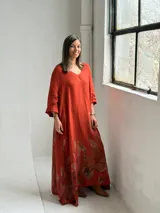 Vestido largo color terracota con escote en V, mangas tres cuartos y diseño amplio, decorado con un estampado floral grande en tono marrón en la parte inferior.