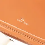 Bandolera de diseño redondeado en color habano, confeccionada en material sintético con acabado liso. Presenta una solapa frontal con cierre de botón imantado y el logo de la marca grabado en el centro. Incluye una correa ancha ajustable de color crema con herrajes plateados.