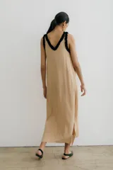 Vestido largo color beige claro, de corte recto y holgado, con escote en V profundo ribeteado en negro con volados. Presenta tirantes finos con el mismo detalle de volado negro.