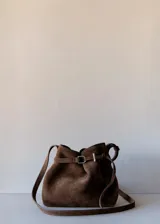 Bolso de mano tipo bandolera confeccionado en piel bovina de color marrón. Cuenta con un diseño estructurado, solapa frontal con detalle de trabilla y hebilla metálica, y asa corta para llevar al hombro o en la mano.