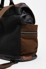 Mochila de estructura blanda confeccionada en nylon color marrón con estampado de cuadros tipo tartán. Presenta bolsillos laterales con cierre, solapa superior con bolsillo frontal y un detalle de cordón elástico en color verde neón.