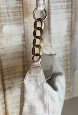 Cartera de hombro color beige con textura tipo cocodrilo, confeccionada en gamuza. Presenta un diseño estructurado con una cadena metálica decorativa en la parte superior del asa y cierre principal.