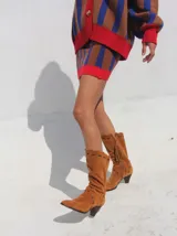 Botas de caña alta confeccionadas en gamuza, con diseño de estilo western. Presentan pespuntes decorativos en cuero, bordado en el empeine y un tirador con flecos en la parte trasera. Cuentan con taco de madera y cierre lateral.