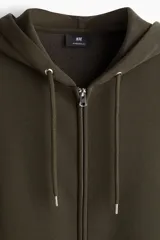 Campera con capucha y cierre frontal, color verde militar oscuro, de tejido pesado con interior cepillado suave. Presenta hombros caídos, cordón ajustable en la capucha y bolsillos canguro delanteros.