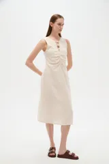 Vestido midi de viscosa color beige, sin mangas y con cuello a la base. Presenta una abertura frontal con botones decorativos y detalles de frunces en el centro del pecho.