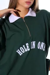 Chomba de piqué color verde con cuello polo blanco y cierre frontal parcial. Presenta un diseño holgado con mangas cortas que incluyen rayas blancas en los puños y un aplique frontal con el texto 'HOLE IN ONE' en color blanco.