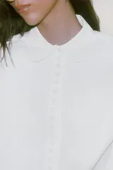 Camisa de popelín color blanco con cuello solapa redondeado y manga larga. Presenta un cierre frontal con botones ocultos y una tapeta decorada con perlas.