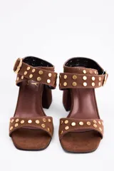 Sandalia de gamuza color marrón con taco cuadrado y punta abierta. Presenta tiras cruzadas en el tobillo con hebilla metálica dorada y detalle de tachas circulares doradas en las tiras y en la parte frontal.