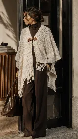 Ruana confeccionada en lana merino color beige, de diseño amplio y envolvente con flecos en el borde inferior y un broche artesanal de cuero en el frente.