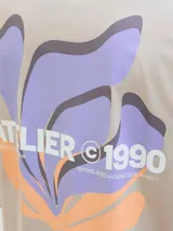 Remera beige de manga corta y cuello redondo con estampado en la espalda con la inscripción "Atelier © 1990" y un diseño abstracto en tonos lila y naranja.