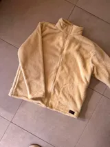 Buzo de poliéster color crema, con textura de sherpa, cuello alto y cierre frontal con cremallera.