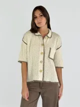 Chaqueta de punto calado en color negro con detalles de bordes en color crudo. Presenta cuello camisero, cierre frontal con botones, mangas cortas y un bolsillo frontal tipo parche.