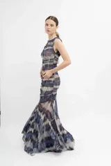 Vestido largo de microtul con estampado tipo batik en tonos azules, grises y blancos. Presenta un diseño sin mangas con cuello redondo y abertura tipo gota en el pecho. El cuerpo está completamente fruncido y forrado, mientras que la falda posee un diseño transparente con terminación en picos que aporta movimiento.