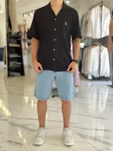 Camisa de manga corta color blanco marfil, confeccionada en tejido texturizado con cuello camisero, cierre frontal de botones y un pequeño bordado oscuro en el pecho.