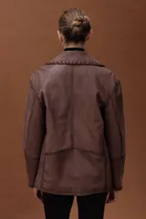 Chaqueta estilo motoquera de cuero ovino color marrón, con diseño oversize. Presenta detalles artesanales de pasamanería en cuello, solapas y bolsillos, cierre frontal cruzado y cierres metálicos en los puños.