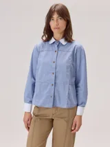 Camisa de manga larga en color azul con textura tipo denim, con cuello y puños en contraste de color blanco. Presenta costuras decorativas horizontales y verticales en el frente, cierre frontal con botones y corte recto.