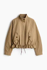 Campera oversized de sarga de algodón color beige, con cuello elevado, cierre frontal oculto y solapa rompevientos. Cuenta con mangas raglán, bolsillos delanteros con solapa y botón, y cordón ajustable en el bajo que frunce la cintura.