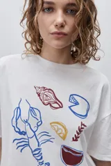 Remera color tostado de corte holgado, con cuello redondo y mangas cortas. Presenta un estampado gráfico frontal de estilo veraniego con ilustraciones de una langosta, conchas, limón, una copa y la inscripción 'Summer Club'.