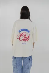 Remera color crema de corte holgado, con cuello redondo y estampado pequeño en el pecho con la frase 'Karma Club' en azul y rojo.
