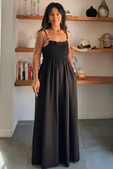 Mono largo de color negro con escote recto y cuerpo superior con fruncido elástico. Presenta breteles finos con detalle de cuentas decorativas y falda amplia con bolsillos laterales.