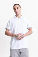 Remera polo blanca de estilo jersey, con fit regular, cuello clásico y tres botones. Presenta un logo estampado en el pecho y detalles de rayas grises en las mangas.
