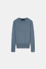 Jersey de punto liso color negro, con cuello redondo y manga larga. Presenta una hilera de cuatro botones dorados decorativos en el puño de cada manga.