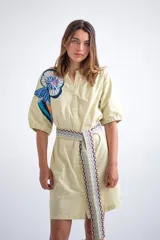 Vestido camisero corto color amarillo pálido con estampado floral en el hombro izquierdo y cinturón de tela con diseño geométrico.