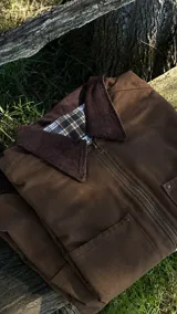 Campera de estilo workwear color chocolate, confeccionada en tela de algodón resistente. Presenta cuello de pana a tono, cierre frontal metálico y bolsillos delanteros. El interior está forrado con tela de franela a cuadros.