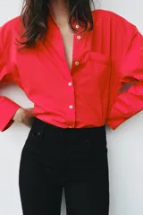 Camisa de popelina roja, de corte oversize, con cuello solapa, manga larga y bajo asimétrico redondeado. Cierre frontal oculto con botones.
