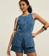 Chaleco de jeans azul de corte alargado, sin mangas, con cuello redondo y cierre frontal de botones dorados. Presenta bolsillos delanteros a la altura de la cadera y un corte que se abre ligeramente en la parte inferior.