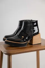 Botineta de cuero negro con diseño combinado en cuero charolado, plataforma y taco cuadrado de madera clara. Presenta cierre lateral y puntera redondeada.