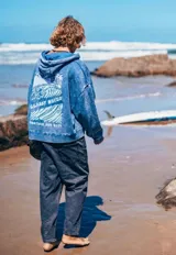 Campera con capucha y cierre frontal doble, confeccionada en tela con efecto lavado azul. Presenta bordado frontal con la frase 'GLASSY WAVES' distribuida sobre los bolsillos tipo canguro.