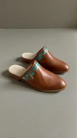 Sandalias tipo mules color marrón terracota, confeccionadas en cuero, con punta redonda, suela plana y detalles pintados a mano en tonos turquesa y dorado con aplique metálico triangular en el lateral.