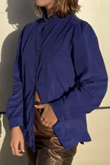 Camisa de pana color verde oliva, con cuello alto con volados, cierre frontal de botones y mangas largas con puños.