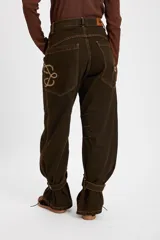 Jean tipo baggy de pierna ancha y recta, color marrón oscuro. Presenta detalles de costuras decorativas, bolsillos laterales redondeados y bolsillos traseros grandes. Incluye tiras ajustables con hebillas metálicas en los tobillos para un efecto fruncido.