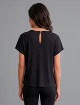 Blusa de modal suave color negro, con mangas cortas y corte holgado. Presenta un escote redondo decorado con una línea de strass y un broche desmontable en forma de moño con strass en el centro. La espalda cuenta con una abertura tipo gota con cierre de botón.