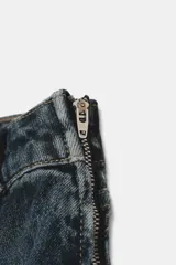 Jeans de mezclilla azul oscuro con lavado desgastado, tiro medio y corte flare ancho. Presenta un detalle de falso cierre con botones metálicos en la parte superior y costuras diagonales marcadas en la parte delantera de las piernas.