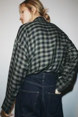 Camisa de mujer con estampado de cuadros vichy en tonos verde oscuro y negro, confeccionada en mezcla de viscosa. Presenta cuello solapa, manga larga con puños, cierre frontal con botones y un detalle de pañuelo integrado en el mismo tejido.