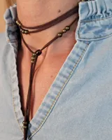 Collar tipo corbatín confeccionado en tiento de cuero marrón, con diseño de doble vuelta y detalle de cuentas metálicas doradas distribuidas a lo largo de las tiras.