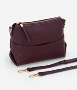 Bolso bandolera pequeño color borgoña, confeccionado en poliuretano con detalle texturizado tejido en la parte inferior. Cuenta con correa transversal ajustable, cierre de cremallera y bolsillo frontal con botón.