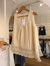Blusa sin mangas de color beige, confeccionada en mezcla de lyocell y lino, con diseño de pliegues en el frente y detalle de encaje tipo puntilla en el ruedo.