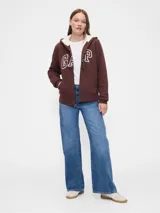 Canguro con cierre frontal, confeccionado en color marrón chocolate, con interior forrado en corderito sintético blanco. Presenta capucha con cordones ajustables, bolsillos tipo canguro y el logo de la marca estampado en el frente en color blanco.