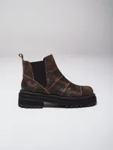 Botas de caña baja con punta cuadrada, confeccionadas en una combinación de cuero liso y gamuza en tonos marrones. Presentan un diseño con múltiples cortes y costuras visibles, cierre elástico lateral y suela de goma dentada tipo track.