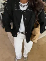 Chaqueta de jean color marrón oscuro, con diseño de hombros caídos y detalle de tachas metálicas en el cuello y la parte superior de las mangas. Presenta cierre frontal con botones metálicos.