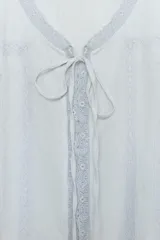 Camisa de gasa color crudo con escote en pico, manga corta abullonada con terminación en volante y cierre frontal con botones ocultos y lazada. Presenta delicados detalles de bordados a tono en toda la prenda.