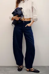 Jean de denim azul oscuro con sobreteñido negro, de tiro medio-alto y corte barrel holgado con piernas amplias que se estrechan en los tobillos, con recortes curvos y bolsillos laterales.