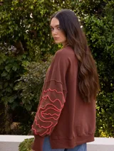 Buzo de tejido de punto color marrón, de corte holgado con tajos laterales. Presenta un bordado artesanal en la manga que simula un contorno de labios en hilo de color coral o naranja neón.