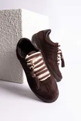 Zapatillas urbanas color tostado con textura geométrica en el cuerpo y detalles de gamuza en la puntera y laterales. Presentan un diseño distintivo con cordones anchos tejidos en tonos tostado y blanco, y un accesorio metálico con borla de gamuza en el empeine.