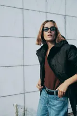 Chaqueta acolchada de color negro con diseño de costuras onduladas, cuello subido y capucha ajustable. Cuenta con cierre frontal de cremallera oculto por una solapa con botones a presión, mangas largas y bolsillos laterales. Tejido con tecnología repelente al agua.